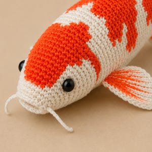 Puede incluir: Un juguete de pez koi tejido a ganchillo a mano con un cuerpo de color crema y parches de color naranja vibrante. El pez tiene dos ojos de cuentas de plástico negro, dos pequeños barbillones que salen de la boca y aletas detalladas.