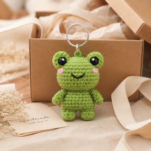 Pode incluir: Um porta-chaves de sapo em croché verde com cabeça redonda, dois olhos de plástico preto, uma boca preta bordada e pequenas bochechas cor-de-rosa. O sapo tem um corpo compacto com braços e pernas curtos, preso a um anel de metal prateado.