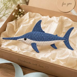 May include: A crocheted swordfish toy featuring a deep blue upper body and a cream-colored underbelly. The fish has a long, pointed bill, a prominent dorsal fin, and a black eye. It is presented inside a brown cardboard gift box with tissue paper.