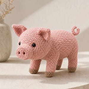 Pode incluir: Uma pequena figura de porco em crochê rosa claro, com um focinho arredondado com duas narinas escuras, duas orelhas pontudas e um pequeno rabo enrolado. O porco tem olhos de contas pretas e quatro patas com detalhes em fio castanho claro nos cascos.