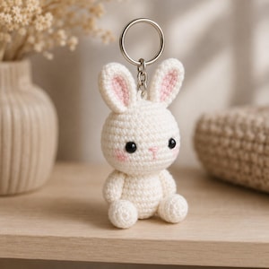 May include: A small, off-white crocheted rabbit keychain featuring two long ears with light pink centers, two black bead eyes, a small pink stitched nose, and two pink blush marks on the cheeks, attached to a silver-toned metal split ring.
