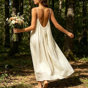 Peut inclure: Une robe longue couleur crème sans manches avec de fines bretelles et un dos nu doté d'un détail à nouer. La robe présente une silhouette ample et fluide et est tenue à côté d'un bouquet de fleurs mélangées blanches, roses et orange.