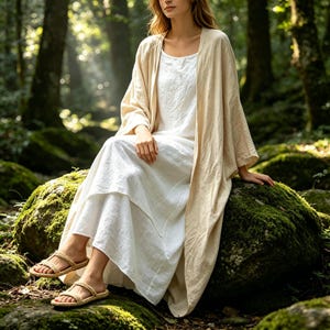 May include: A long-sleeved beige open-front robe worn over a floor-length white sleeveless dress. The outfit is paired with tan woven rope sandals featuring two straps across the top of the foot and a simple ankle strap design.