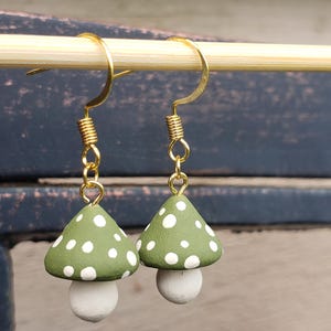 May include: A pair of dangle earrings featuring mushroom-shaped charms. Each charm has a matte olive green cap with white polka dots and a solid white stem. The charms are attached to gold-toned metal fishhook ear wires with a small coiled detail.