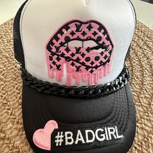 May include: A black and white trucker hat featuring a pink dripping lip patch with a black monogram pattern. The hat includes a thick black plastic chain across the brim, a pink heart patch, and a black rectangular patch with the text #BADGIRL.