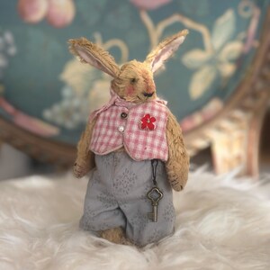 May include: A plush rabbit toy with tan fur, wearing a pink and white checkered vest with a small red flower accent and a single pearl button. The rabbit is dressed in grey patterned trousers and has a metal skeleton key hanging from its waist.