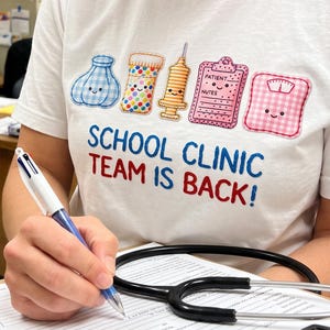 May include: A white t-shirt featuring a graphic of a blue ice pack, a pill bottle, a syringe, a pink patient notes clipboard, and a pink scale, with the text SCHOOL CLINIC TEAM IS BACK! in blue and red letters, worn while holding a clipboard.