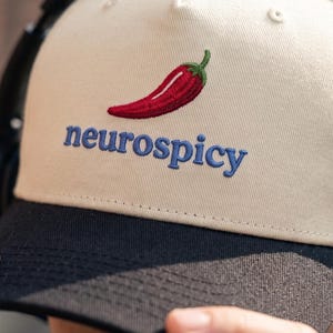 Op de afbeelding: Een beige honkbalpet met een zwarte klep, voorzien van een geborduurd rood chilipeper-icoon boven de blauwe tekst "neurospicy" in het midden van het voorpaneel. De pet heeft een gestructureerde kroon en een platte klep met zichtbare stiksels.