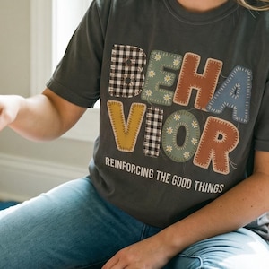 May include: A charcoal gray short-sleeve t-shirt featuring the word BEHAVIOR in large, multi-patterned, stitched-style letters. Below the word, the text reads REINFORCING THE GOOD THINGS in white capital letters. The shirt has a crew neckline.