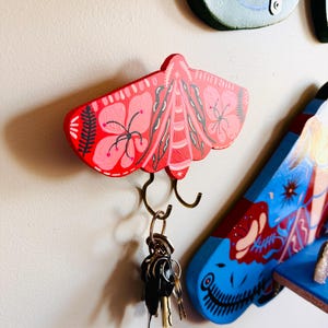 May include: A red wooden wall-mounted hook shaped like a moth with pink floral patterns and dark grey fern-like details. Two curved metal hooks extend from the bottom, holding a metal keyring with several silver and black keys attached to it.