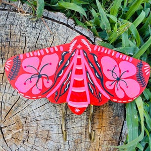 May include: A wall-mounted hook featuring a hand-painted wooden moth in vibrant shades of red and pink. The moth wings display symmetrical floral patterns in light pink with black accents, while two curved brass-toned metal hooks extend downward.