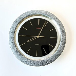 May include: A round wall clock featuring a black face with thin gold-tone hour markers and hands. The clock has a wide, textured silver glitter frame and a white inner border. The word Quartz is printed in white at the bottom of the clock face.