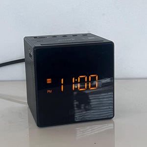 May include: A black cube-shaped Sony digital alarm clock with a glossy front display showing the time 11:00 in bright orange LED digits. The device features a PM indicator and a radio signal icon on the left side of the digital clock face.