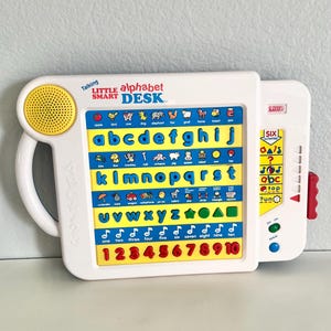 May include: A white VTech Talking Little Smart Alphabet Desk with a yellow speaker, blue letter keys, and red number buttons. The device features rows of colorful icons, shapes, and numbers, with a side panel containing six activity buttons.