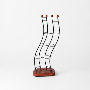 May include: A vertical wine rack featuring a wavy black metal frame with four circular bottle holders. The structure is supported by a rectangular wooden base and topped with four small wooden spheres at the end of each metal wire rod.