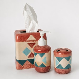 May include: A ceramic bathroom set consisting of a rectangular tissue box cover, a cylindrical soap dispenser with a white pump, and a cylindrical toothbrush holder. Each piece features a terracotta, beige, and teal geometric triangle pattern.