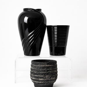 May include: A collection of three black vessels including a glossy vase with a swirling side design, a glossy cylindrical container with horizontal ridges, and a matte, textured, barrel-shaped pot with horizontal bands and a weathered finish.