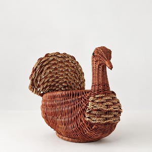 May include: A woven wicker basket shaped like a turkey, featuring a reddish-brown body with a lighter tan woven tail fan and wing accents. The basket has a long neck, a curved beak, and a small dark eye, constructed from natural plant fibers.