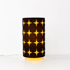May include: A cylindrical table lamp featuring a black fabric shade with a repeating pattern of yellow four-pointed stars. The lamp emits a warm glow from within, illuminating the star shapes, and stands on small, thin, silver-colored metal legs.