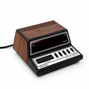 May include: A vintage Spartus digital alarm clock featuring a dark wood grain finish on the top and sides, a black plastic base, and a dark display screen. The control panel includes a large snooze button and switches labeled Hour, Min, Time Set, and Alarm.