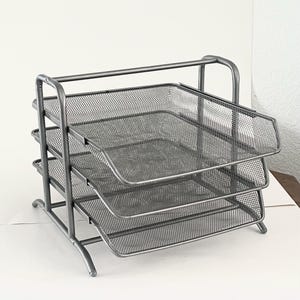 May include: A silver-colored metal desk organizer featuring three stacked horizontal trays made of fine wire mesh. The structure is supported by a sturdy tubular frame with curved side supports, providing a tiered system for sorting documents.