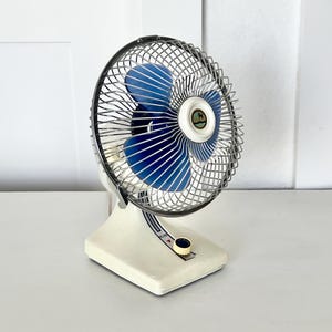 Könnte beinhalten: Ein elektrischer Tischventilator im Vintage-Stil mit einem cremefarbenen Kunststofffuß und einem runden Metallgitter. Der Ventilator hat vier durchscheinende blaue Kunststoffflügel und eine weiße Kappe in der Mitte mit einem kleinen Logo.