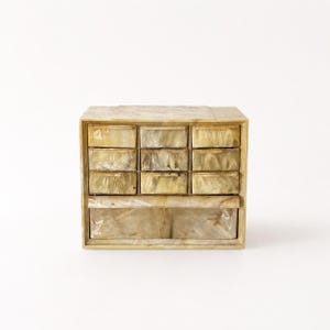 May include: A rectangular storage chest featuring a light beige and cream marbled resin finish. The unit includes nine small square drawers arranged in three rows of three, positioned above one wide, horizontal drawer at the base.