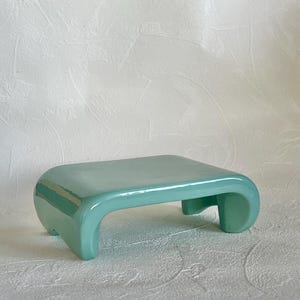 May include: A small, rectangular, light mint green ceramic display stand with a smooth, glossy finish. The stand features a flat top surface and four curved, integrated legs that extend from the corners, creating a low-profile, elevated platform.