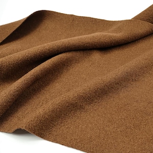 May include: A piece of brown felt fabric with a textured, matte surface. The material is draped to show soft folds and curves, highlighting the consistent medium-brown color and the dense, fibrous quality of the textile across the entire surface.