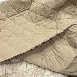 Può includere: Un tessuto trapuntato beige caratterizzato da un motivo a cuciture ondulate ripetute. Il materiale presenta una finitura opaca e morbida sullo strato superiore e una fodera leggermente più liscia e chiara visibile sul bordo piegato, mostrando la struttura.