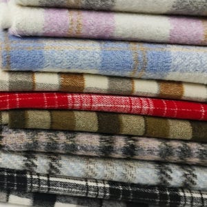 Op de afbeelding: Een stapel gevouwen stoffen met diverse ruit- en plaidpatronen. De textielstukken hebben een zachte, geborstelde textuur met kleurcombinaties waaronder lavendel, lichtblauw, beige, rood, olijfgroen, grijs, en zwart met wit.