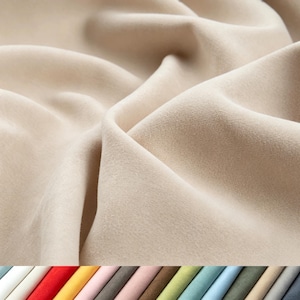 Peut inclure: Un tissu beige à la texture douce et mate rappelant le daim, présentant des plis profonds et des ombres marquées. Sous le tissu principal, une rangée horizontale de petits échantillons rectangulaires affiche une variété de couleurs, dont le blanc, le rouge, le jaune, le rose, le gris, le vert et le bleu.
