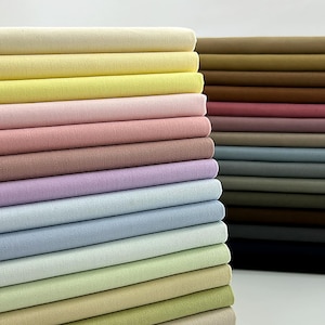 Puede incluir: Una pila de piezas de tela dobladas en una variedad de colores sólidos, incluyendo tonos crema, amarillo, rosa, lavanda, azul claro y verde menta, dispuestas en una pila vertical ordenada junto a otra pila de telas en tonos tierra oscuros.
