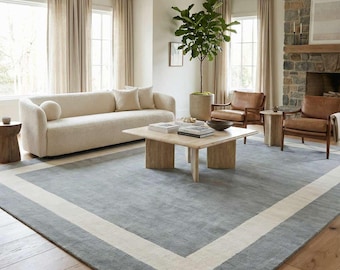May include: A large rectangular area rug featuring a solid slate blue center field framed by a wide, contrasting cream-colored border. The rug is crafted with a low-pile texture and is positioned on a light-toned hardwood floor in a living room.