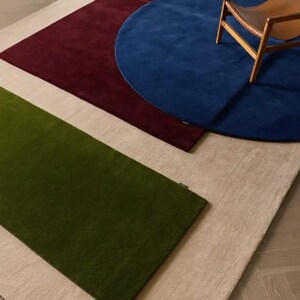 May include: A collection of layered rugs featuring a large rectangular beige base, a rectangular olive green rug, a rectangular deep burgundy rug, and a circular royal blue rug. A wooden chair with a tan seat is positioned on the blue rug.