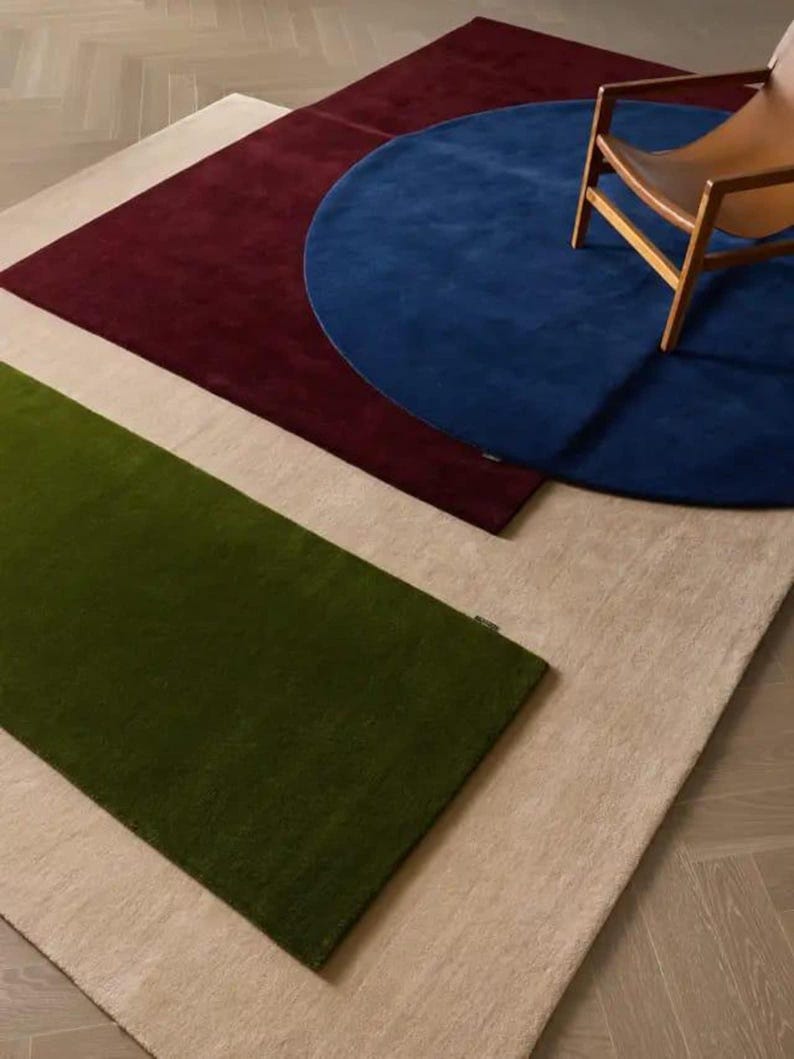 May include: A collection of layered rugs featuring a large rectangular beige base, a rectangular olive green rug, a rectangular deep burgundy rug, and a circular royal blue rug. A wooden chair with a tan seat is positioned on the blue rug.