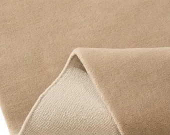 May include: A solid beige area rug with a soft, dense pile texture. The rug is folded over at one corner, revealing a lighter, woven backing material underneath the plush, uniform surface of the main carpet fabric.