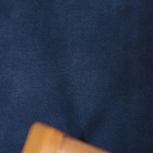 May include: A dark navy blue fabric with a soft, matte texture positioned behind a light brown wooden frame edge. The fabric appears uniform in color and density, providing a solid, dark backdrop to the partially visible wooden structure in the foreground.