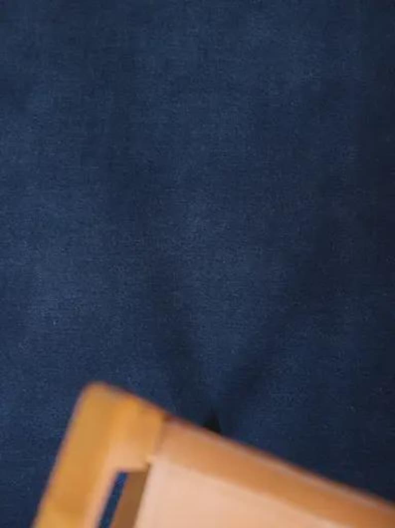 May include: A dark navy blue fabric with a soft, matte texture positioned behind a light brown wooden frame edge. The fabric appears uniform in color and density, providing a solid, dark backdrop to the partially visible wooden structure in the foreground.