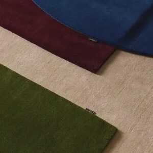 May include: Three rectangular rugs layered over one another, featuring a deep blue rug on top, a dark burgundy rug in the middle, and a textured olive green rug at the bottom, all resting on a light beige surface with a small brand tag on each.