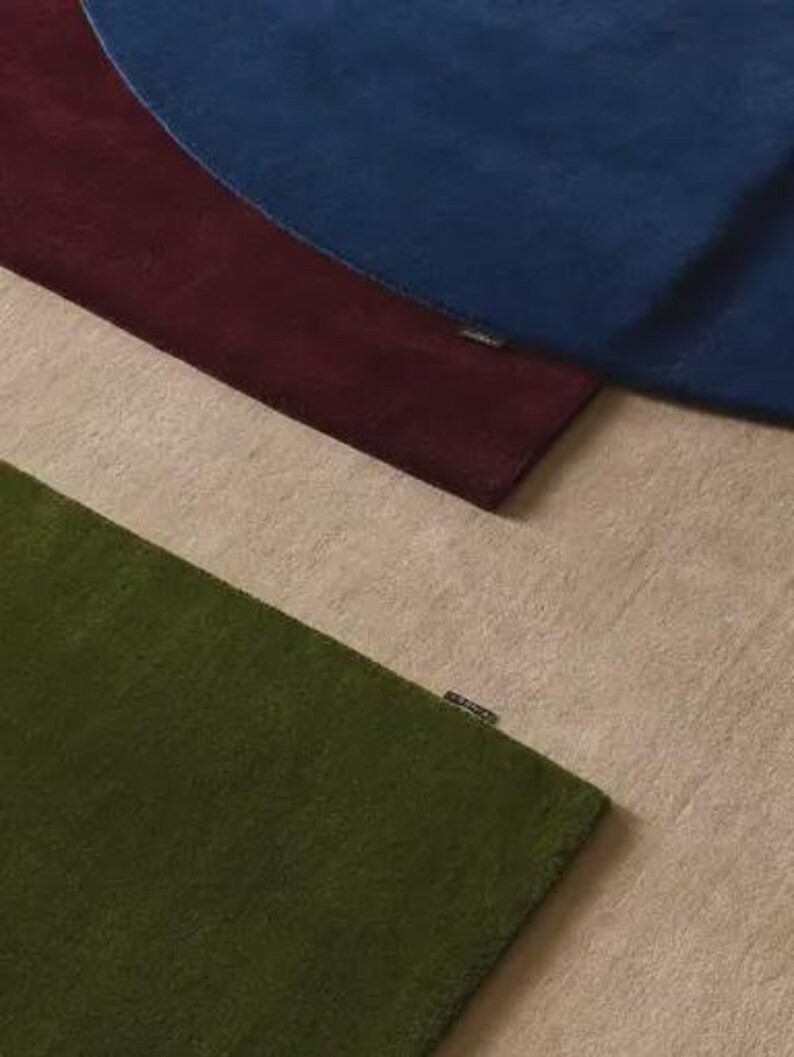 May include: Three rectangular rugs layered over one another, featuring a deep blue rug on top, a dark burgundy rug in the middle, and a textured olive green rug at the bottom, all resting on a light beige surface with a small brand tag on each.