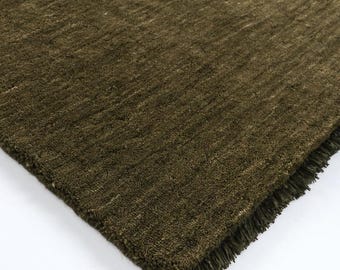 May include: A rectangular area rug in a deep olive green color with a dense, textured pile surface. The rug features a straight, finished edge along one side and a short, matching olive green fringe trim along the adjacent perpendicular edge.