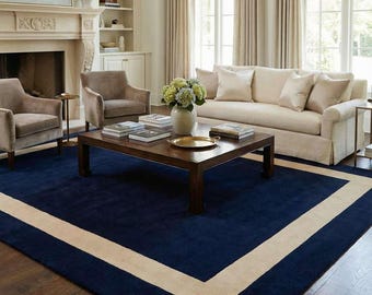 May include: A large rectangular area rug featuring a deep navy blue center field framed by a wide, solid beige border. The rug is positioned on a dark wood floor, providing a bold contrast to the surrounding neutral-toned living room furniture.