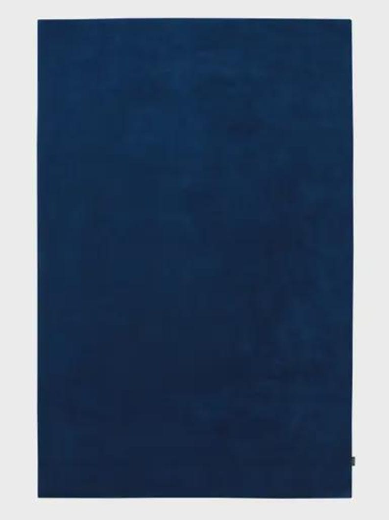 May include: A rectangular textile piece in a solid, deep navy blue color. The fabric features a consistent, soft-textured surface with a subtle matte finish. A small, dark rectangular tag is visible on the bottom right edge of the item.