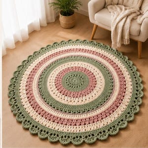 May include: A round, crocheted rug featuring concentric circles in alternating shades of sage green, dusty rose, and cream. The rug has a textured, intricate stitch pattern throughout and is finished with a decorative scalloped edge border.