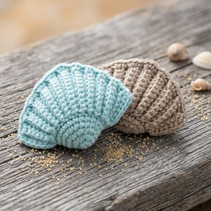 May include: Two crocheted seashell ornaments, one in light blue and one in beige, featuring a ribbed texture and a fan-like shape. The items are crafted with yarn and display a detailed stitch pattern that mimics the appearance of natural sea shells.