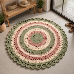 May include: A round, crocheted rug featuring concentric circles in shades of sage green, dusty rose, and cream. The rug has a textured, stitch-patterned surface and a decorative, scalloped edge that creates a finished, circular border design.