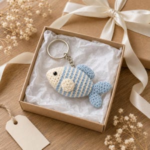 May include: A crochet fish keychain featuring alternating light blue and cream stripes, a small round cream fin, and a solid light blue tail. The fish has a single black bead eye and is attached to a silver-toned metal split ring key holder.