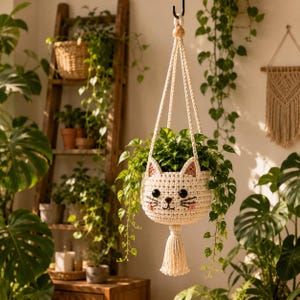 Peut inclure: Une jardinière suspendue en crochet de couleur crème en forme de visage de chat avec des yeux en boutons noirs, un nez et une bouche brodés, des joues roses et des oreilles pointues. Elle est suspendue par des cordes tressées et un pompon.