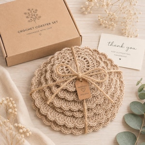 May include: A set of round, beige crochet coasters tied with twine and a small tag reading "HANDMADE WITH LOVE". The set includes a brown cardboard box labeled "CROCHET COASTER SET HANDMADE WITH LOVE" and a card reading "thank you".